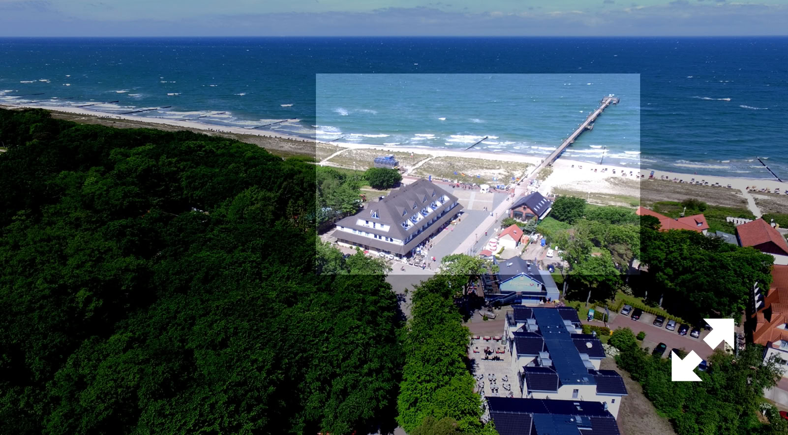 Luftaufnahme einer Küstenlandschaft mit einem Pier, einem großen Gebäude und mehreren kleineren Gebäuden, umgeben von Bäumen und dem Meer im Hintergrund.