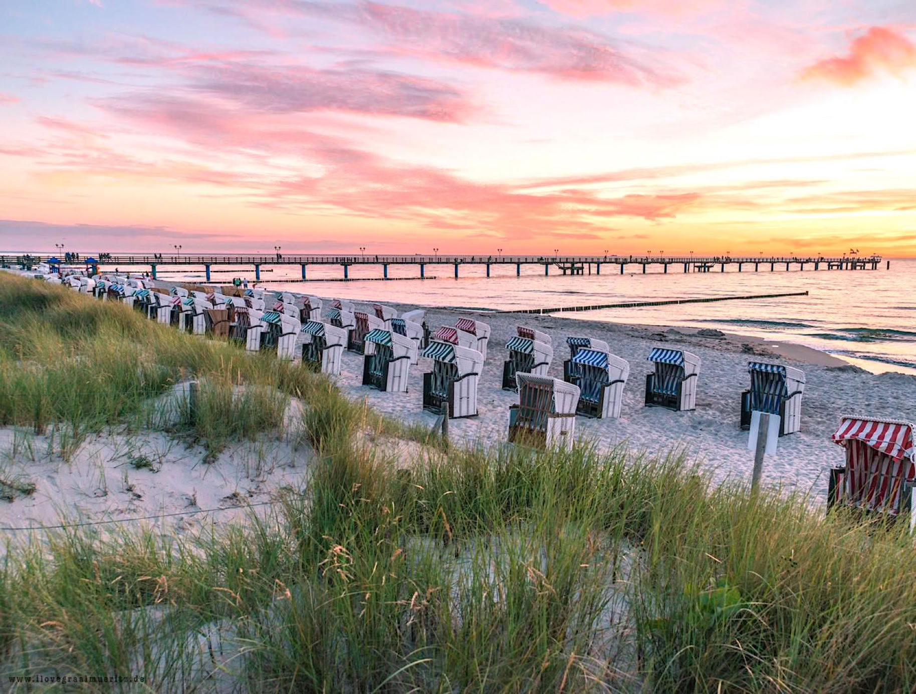 Strand mit Strandkörben im Vordergrund, Grasbüschel und ein langer Steg im Hintergrund, Sonnenuntergang am Horizont.